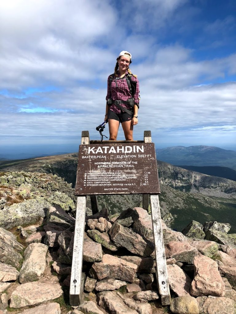 Heidi Nisbett wears hiking gear and stands on a pile of boulders at the top of a mountain behind a sign reading "Katahdin", signifying the completion of her thru-hike of the Appalachian Trail.